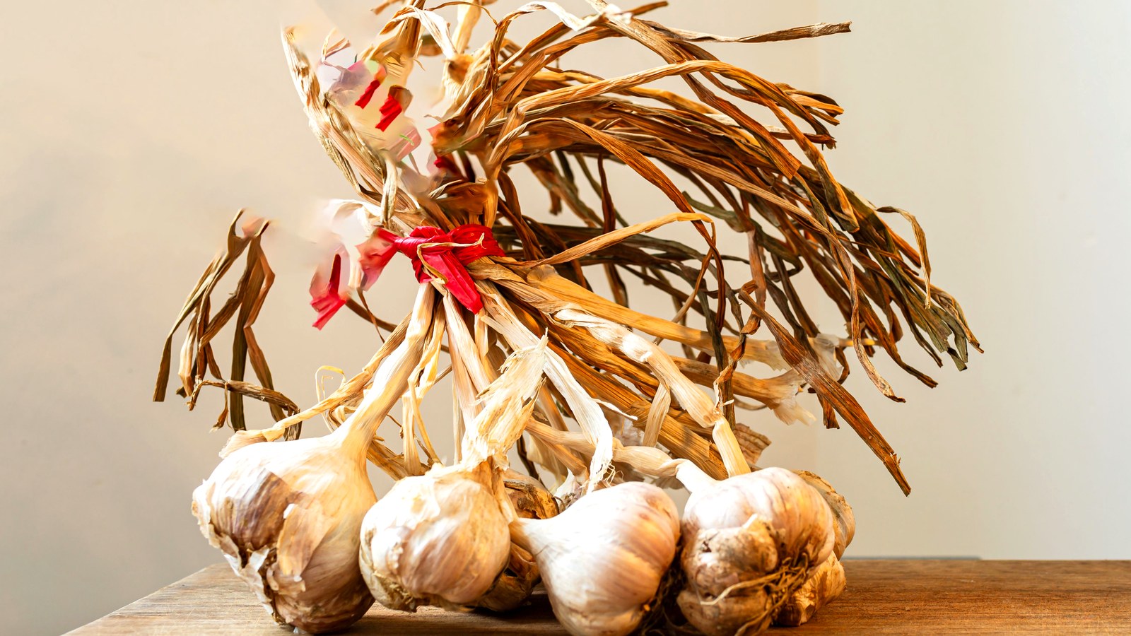 Bundle of cured garlic bulbs tied with red string, lying on a wooden table against a pale wall.