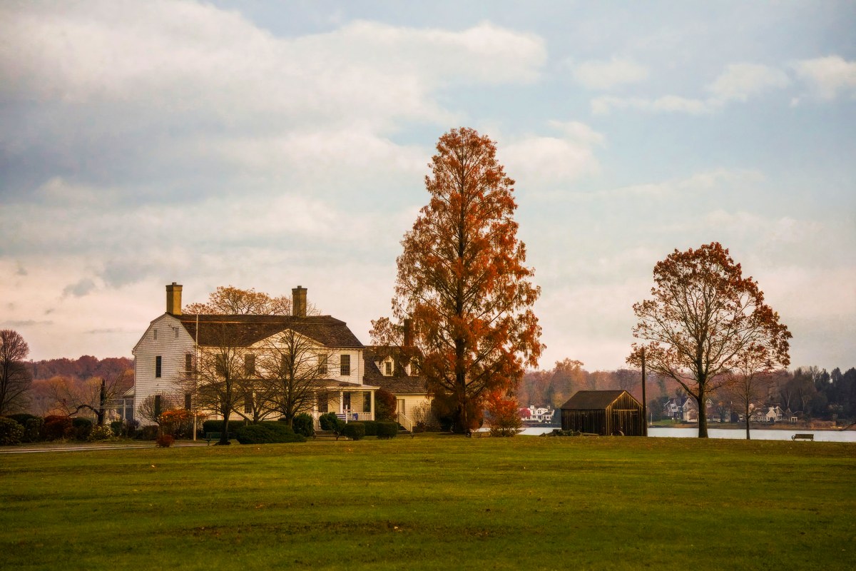 A wide green lawn before a white country house, with tall trees in orange fall color.