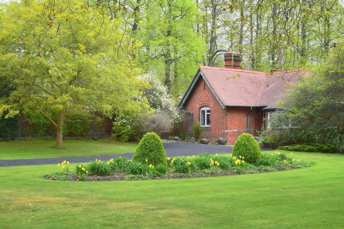 A green lawn around a round planted bed of shrubs and flowers in front of a brick cottage.