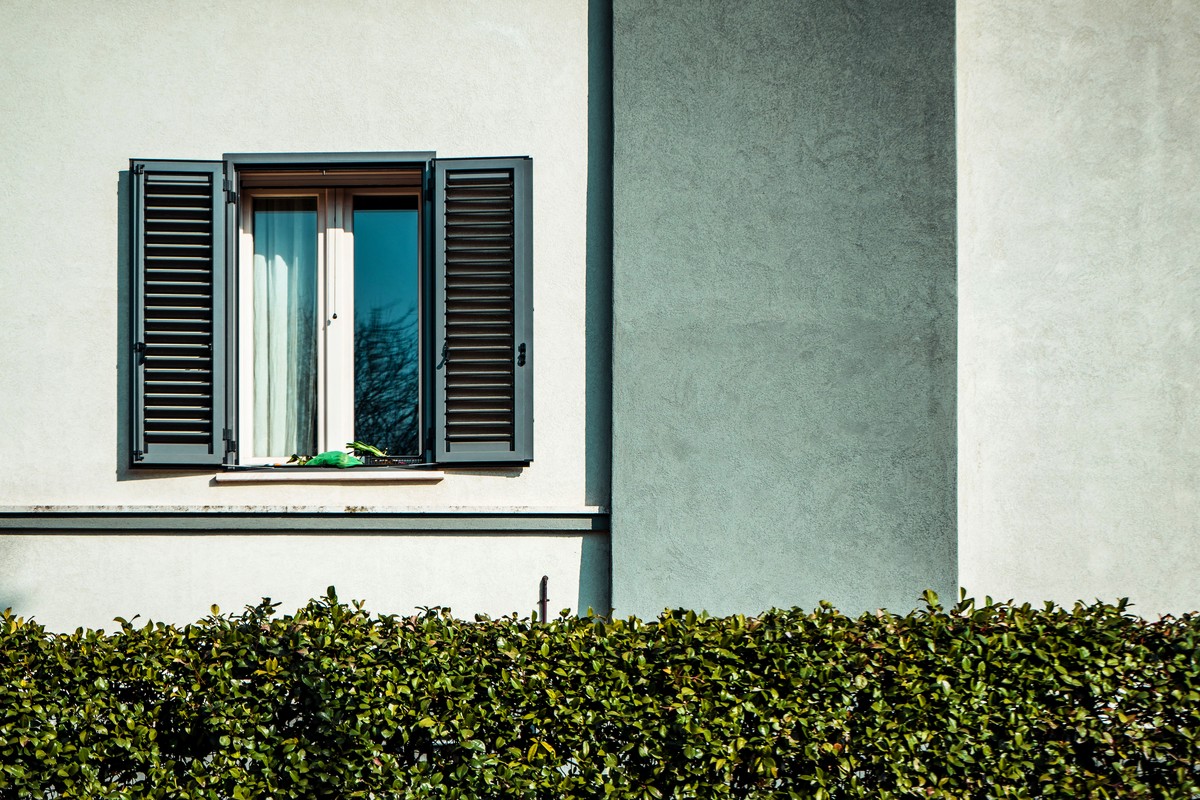 A shuttered window sits just above a clipped hedge, the spot where a thrown rock could crack glass.